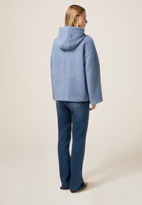 Sudadera azul claro con capucha y de corte oversized en punto texturizado, combinada con jeans de mezclilla oscuros y de corte acampanado. El conjunto presenta detalles de diseño minimalistas.