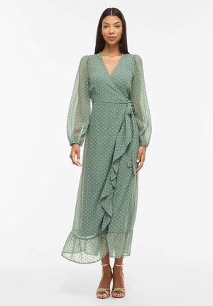 Kobieta w zielonej sukience kopertowej z tiulowymi długimi rękawami, w groszki, z falbanami, w złotych sandałkach na paski, stojąca na białym tle.