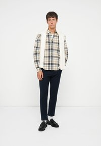 Een geruite shirt in beige, marineblauw en lichtbruin, gecombineerd met marineblauwe broek. Een witte gebreide trui over de schouders; zwarte schoenen.