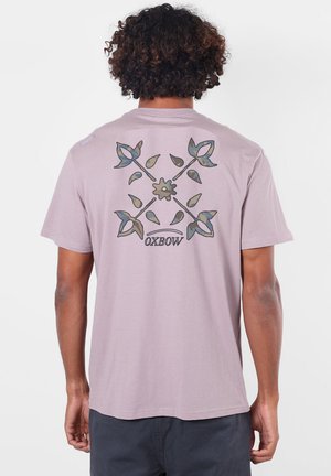 Personne aux cheveux bouclés portant un t-shirt violet clair avec un motif de feuilles bleu-vert croisées et de fleurs avec le texte "OXBOW" dans le dos.