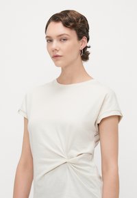 Lys beige t-skjorte med kort erm, rund hals og vridd detalj foran. Myk bomullsmateriale med rullede ermer.