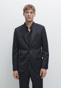 Dunkler Nadelstreifenanzug mit einer maßgeschneiderten Jacke mit zwei Knöpfen, kragenförmigen Revers und zwei Fronttaschen. Dazu ein passendes Hemd.