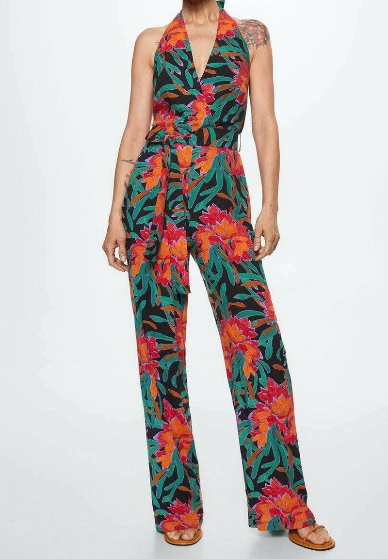 Combinaison sans manches à col halter avec jambes larges, fond noir, grand imprimé floral orange et rouge, feuilles vertes et ceinture à la taille, portée avec des sandales.