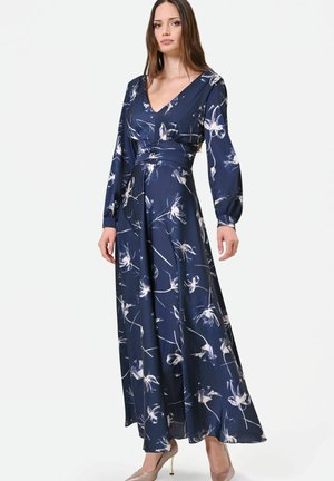 Femme portant une longue robe fleurie bleu marine avec manches longues, un décolleté en V, une taille cintrée et des chaussures à talons beige, debout sur fond blanc.