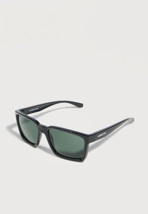 Lunettes de soleil Arnette à monture rectangulaire noire avec des verres verts foncés reposant sur une surface plate gris clair.