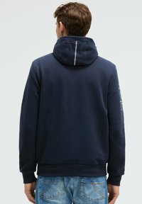 Sudadera azul marino con capucha ajustable, puños y bajo acanalados. Presenta una línea blanca contrastante en la capucha y detalles impresos en la manga.
