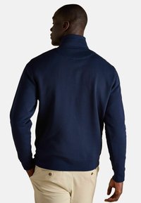 Tumši zils džemperis ar augstu apkakli, ribotām piedurknēm un gludu tekstūru, rādīts no mugurpuses uz modeli, kurš valkā gaišas bikses.