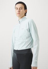 Person som har på seg en lys blå langermet treningsgenser med kvart glidelås og svarte tettsittende leggings mot en ensfarget bakgrunn.