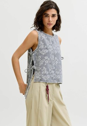 Žena s krátkymi tmavými vlasmi, ktorá má na sebe sivý kvetovaný top bez rukávov so zaväzovaním po stranách a béžové plisované nohavice s červeným príveskom na kľúče.