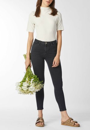 Vrouw in een wit kort mouw coltruitje, zwarte hooggetailleerde skinny jeans en bruine sandalen die een boeket witte bloemen vasthoudt tegen een witte achtergrond.