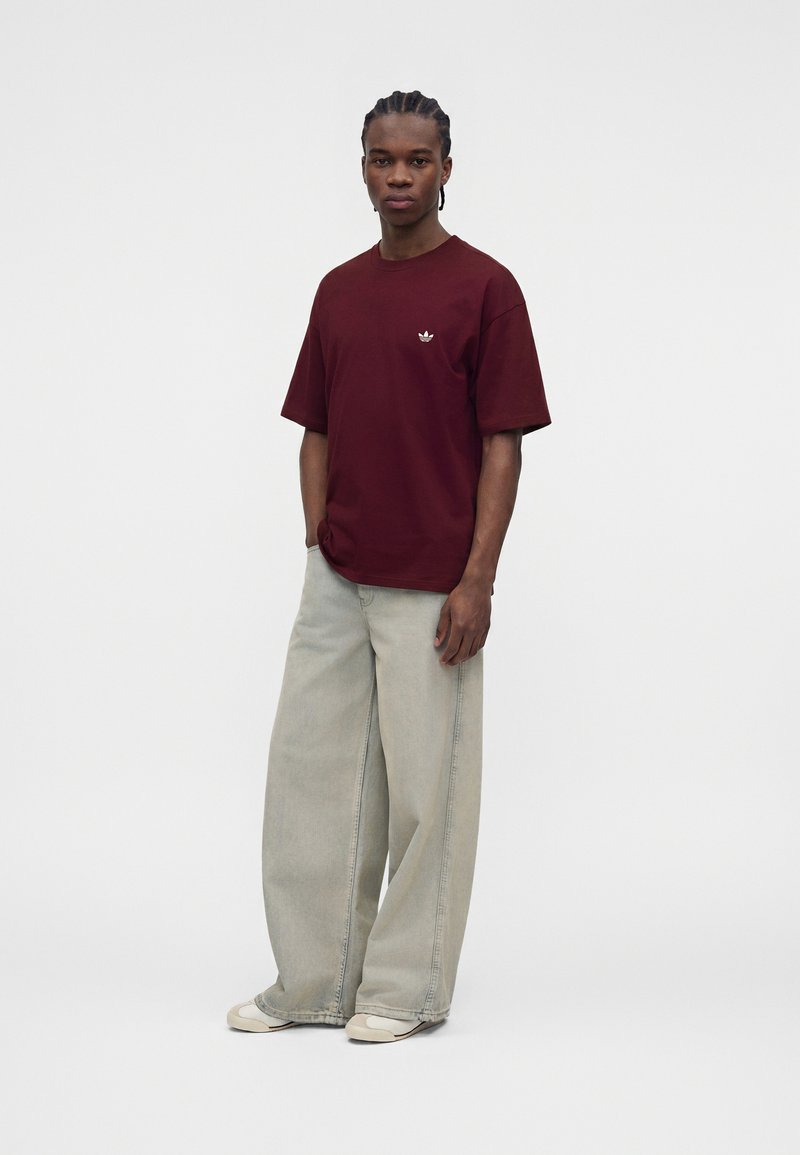 Mann trägt ein burgunderrotes Adidas-T-Shirt, breite hellgraue Jeans und weiße Sneakers, steht mit den Händen in den Taschen vor einem einfarbigen Hintergrund.