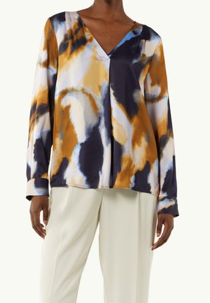 Blouse à manches longues avec col en V et motif abstrait moutarde, marine et blanc, associée à un pantalon crème taille haute sur une personne debout.