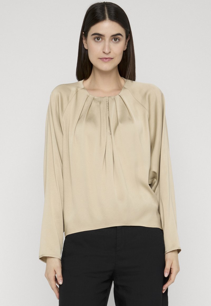 Femme aux cheveux noirs et lisses portant un chemisier beige à manches longues avec des plis près de l'encolure et un pantalon noir, debout devant un fond gris.