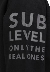 Zwarte sweatshirt met grote, vetgedrukte, grijze tekst "SUB LEVEL ONLY THE REAL ONES" op de voorkant gedrukt. Zachte stoftextuur.