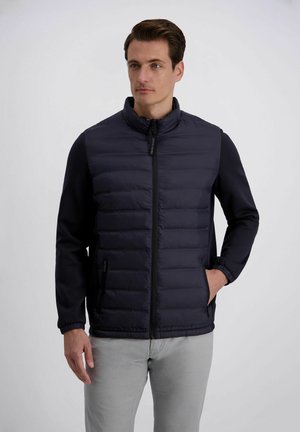 Mann trägt eine schwarze gesteppte Jacke mit Reißverschluss und langen Ärmeln sowie hellgraue Hosen, steht vor einem schlichten weißen Hintergrund.