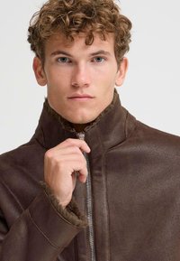 Braune Lederjacke mit hohem Kragen, ausgestattet mit einem Reißverschluss und weichen, mit Fell gefütterten Manschetten. Das Material hat eine strukturierte Oberfläche.