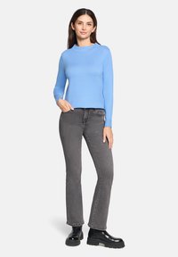 Kobieta ubrana w jasnoniebieski sweter z długim rękawem, szare dopasowane jeansy oraz czarne masywne botki za kostkę, stojąca z jedną ręką w kieszeni.