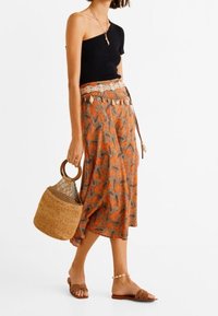 Musta one-shoulder toppi yhdistettynä virtaavaan oranssiin kuviolliseen hameeseen, kudottuun olkikassiin ja ruskeisiin liukuviin sandaaleihin, joissa on teksturoitu hihna.