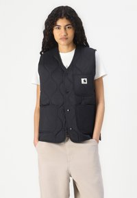 Gilet trapuntato nero con scollatura a V, dotato di due tasche frontali e un piccolo logo. Indossato sopra una maglietta bianca e pantaloni beige.