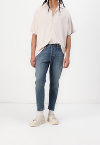 Camicia button-up a maniche corte color beige chiaro, jeans in denim blu, sneakers color crema; vestibilità rilassata; texture morbida, design casual, leggero sbiadimento nei jeans.