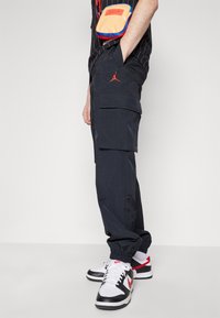 Svarta cargobyxor i lätt material med synliga sidofickor och en röd Jordan-logotyp. Matching med svarta och vita sneakers.