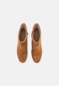 Stivaletti alla caviglia in suede marrone con una texture liscia, chiusura con zip laterale e design a punta rotonda. L'interno mostra il marchio.