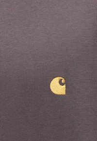 Καφέ ύφασμα με χρυσό κεντημένο λογότυπο Carhartt στο κέντρο της δεξιάς πλευράς.