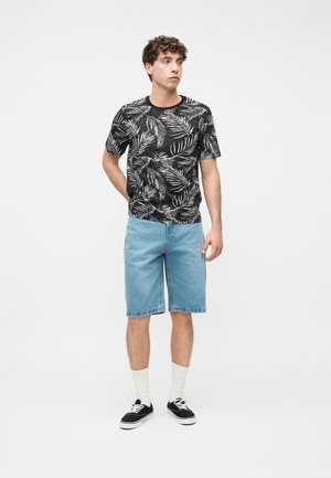 Jeune homme debout, mains derrière le dos, portant une chemise noire et blanche à motif de feuilles, un short en denim bleu clair, des chaussettes blanches et des baskets noires.