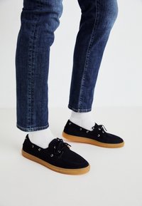 Schwarze Wildleder-Bootsschuhe mit braunen Gummisohlen, ausgestattet mit weißen Ösen und Schnürsenkeln, kombiniert mit dunkelblauen Jeans und weißen Socken.