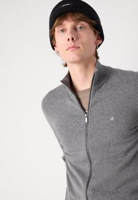 Un sweat à fermeture éclair gris avec un col côtelé, présentant un petit logo sur la gauche. Le modèle porte un bonnet en tricot noir et une chemise beige en dessous.