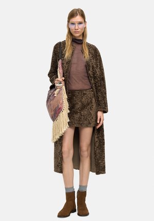 Kobieta w brązowym, teksturowanym płaszczu i spódnicy, przezroczystej brązowej bluzce, okularach z niebieskim odcieniem, brązowych botkach, szarych skarpetkach, niosąca torbę typu tote z frędzlami.