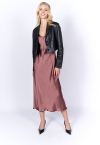Giacca in pelle nera corta indossata su un vestito di raso malva, caratterizzato da una superficie liscia e una silhouette aderente. La modella indossa tacchi a punta neri.
