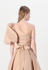 One-shoulder, beige dress made of textured fabric, featuring a large puff sleeve, ruched bodice, and a tied bow detail at the back.