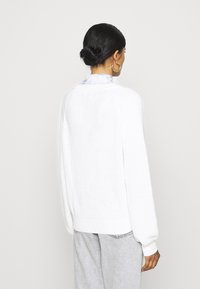 Suéter blanco de punto con puños y dobladillo acanalados, que presenta un corte holgado y una tela texturizada, combinado con pantalones gris claro.