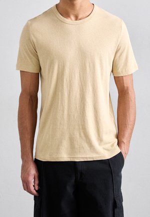 Beige cotton t-shirt with a crew neck and short sleeves. Soft texture, minimal stitching, and a regular fit. Paired with black cargo shorts.