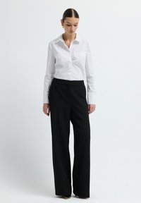 Vrouw in een wit overhemd met knopen en zwarte wijde broek, staand tegen een effen lichte achtergrond, naar beneden kijkend.