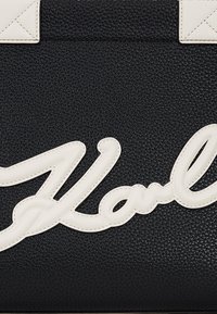 Geantă de umăr din piele neagră cu un finisaj texturat, logo alb semnat în litere reliefate și accente crema contrastante pe mânere.