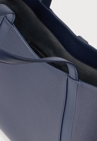 Tmavomodrá kožená taška cez rameno s textúrovaným vonkajškom, hladkými remienkami a vnútorným vreckom na zips, ponúkajúca dve možnosti nosenia.