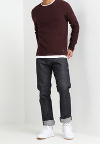 Bordeauxroter Strickpullover mit strukturiertem Muster, getragen über einem weißen Hemd, kombiniert mit dunklen, hochgekrempelten Jeans und weißen Sneakern.