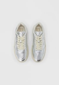 Hopeanväriset urheilukengät, joissa on rei'itetty muotoilu, beiget nyörit ja kumipohjat. Sileä metallinhohtoinen päällinen ja hillitty logoyksityiskohta sivulla.