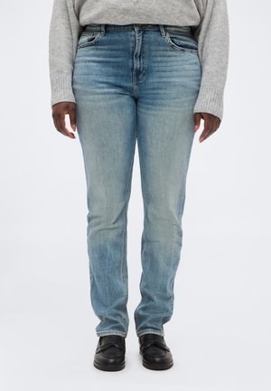 Oseba, oblečena v svetlo modre kavbojke, sivi pleteni pulover in črne natikače, stoji ob belem ozadju.