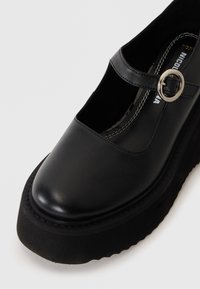 Μαύρο δερμάτινο παπούτσι Mary Jane με χοντρή σόλα και ασημένιο λουράκι με αγκράφα σε λευκό φόντο.