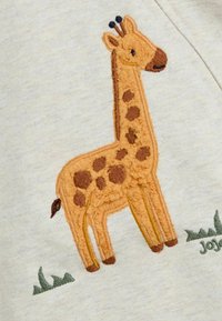 Design de girafe brodé en tan et marron sur un tissu beige clair, avec des taches, des antennes et des accents d'herbe verte en dessous.