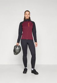 Bordeauxrode en zwarte fietsjacket met een hoge kraag, oranje ritssluitingdetail en nauwsluitende zwarte legging. Zwarte helm in de linkerhand gehouden.