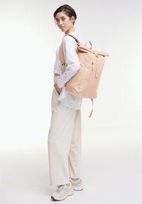 Beige roltop rugzak met verstelbare banden, ritsvak en een soepele textiele afwerking. Model draagt een wit shirt en lichte broek.