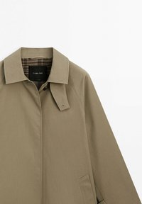 Khaki trenchcoat med kraveklap og knap, med ternet foer indvendigt og sidelomme, lagt fladt ud på hvid baggrund.