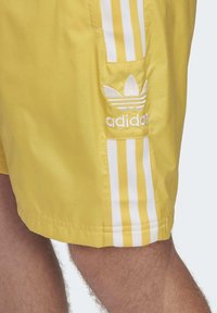Gula shorts i lätt material med vita vertikala ränder och en broderad vit Adidas-logotyp på vänster ficka.