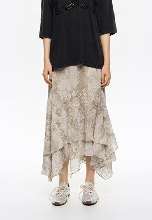 Luzna, warstwowa spódnica midi w jasnym beżu z subtelnym wzorem w węża; zestawiona z luźną czarną bluzką i metalicznymi sneakersami.