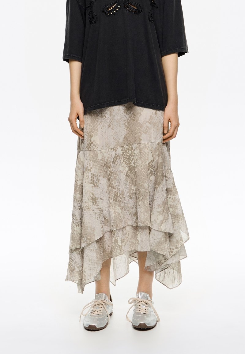 Falda midi fluida y con capas en beige claro con un sutil estampado de serpiente; combinada con una blusa negra holgada y zapatillas metálicas.