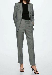 Costume gris sur mesure avec un blazer cintré et un pantalon slim. Le tissu présente un motif chevrons subtil, porté avec un haut noir et des talons à brides.